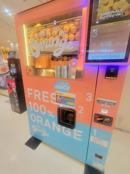 Fresh 100%オレンジと書かれた自動販売機。中にたくさんの生オレンジ果実が見える。