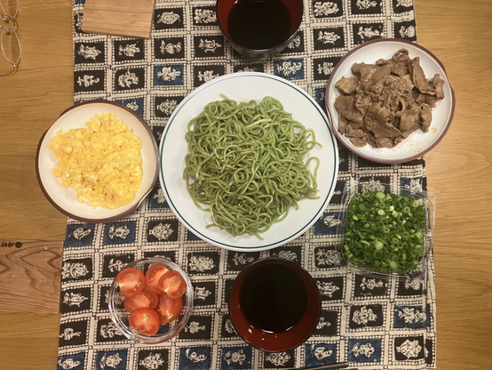 食卓を上から撮影。真ん中に緑色の茶そば、周辺にいりたまご、肉、ネギ、つけダレなどがある。