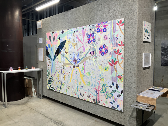 壁にカラフルで大きな絵がかかっている。絵は切り絵で、花などが表されている。台に小さなオブジェが4点並んでいる。